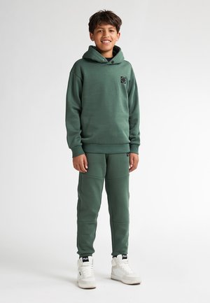 Zelená mikina s kapucí a logem, spárovaná s odpovídajícími tepláky s hladkou texturou. Bílé vysoké tenisky dotvářejí outfit.
