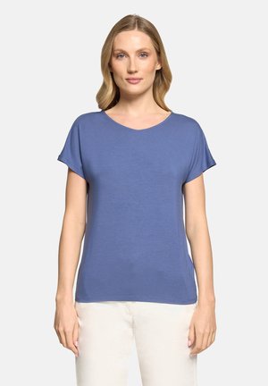 Femme aux cheveux blonds mi-longs portant un haut bleu à manches courtes et un pantalon blanc, debout devant un fond clair uni.