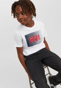 Balta kokvilnas T-krekls ar zilu un sarkanu grafiku, kurā ir "JACK & JONES" un vertikālās svītras, savienots ar melnajām džinsām un sporta apaviem.