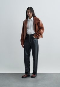 Καφέ δερμάτινο μπουφάν με μεγάλο γιακά, λευκό πουκάμισο, μαύρο παντελόνι με φαρδιά πόδια και μπορντό παπούτσια. Λείο υλικό, με αντίθετες ραφές.