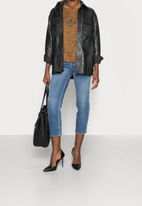 Černá kožená košile-jacket přes zlatý svetr, spárovaná se světle modrými zkrácenými džíny a černými botami na podpatku s špičatou špičkou. V jedné ruce drží černou tašku.