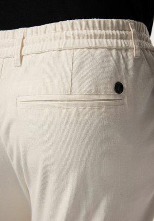 Pantalons couleur crème avec une texture côtelée, dotés d'une taille élastique et d'une seule poche arrière avec un détail en métal noir.