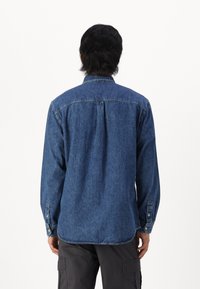 Camisa de mezclilla en azul oscuro con cuello tipo button-down, mangas largas y diseño en el yugo trasero. Presenta puños con detalles de botones.