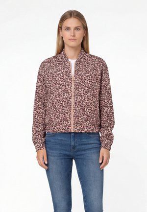 Giovane donna in piedi, indossa una giacca con cerniera rosa e marrone a stampa leopardata e jeans blu su uno sfondo bianco uniforme.