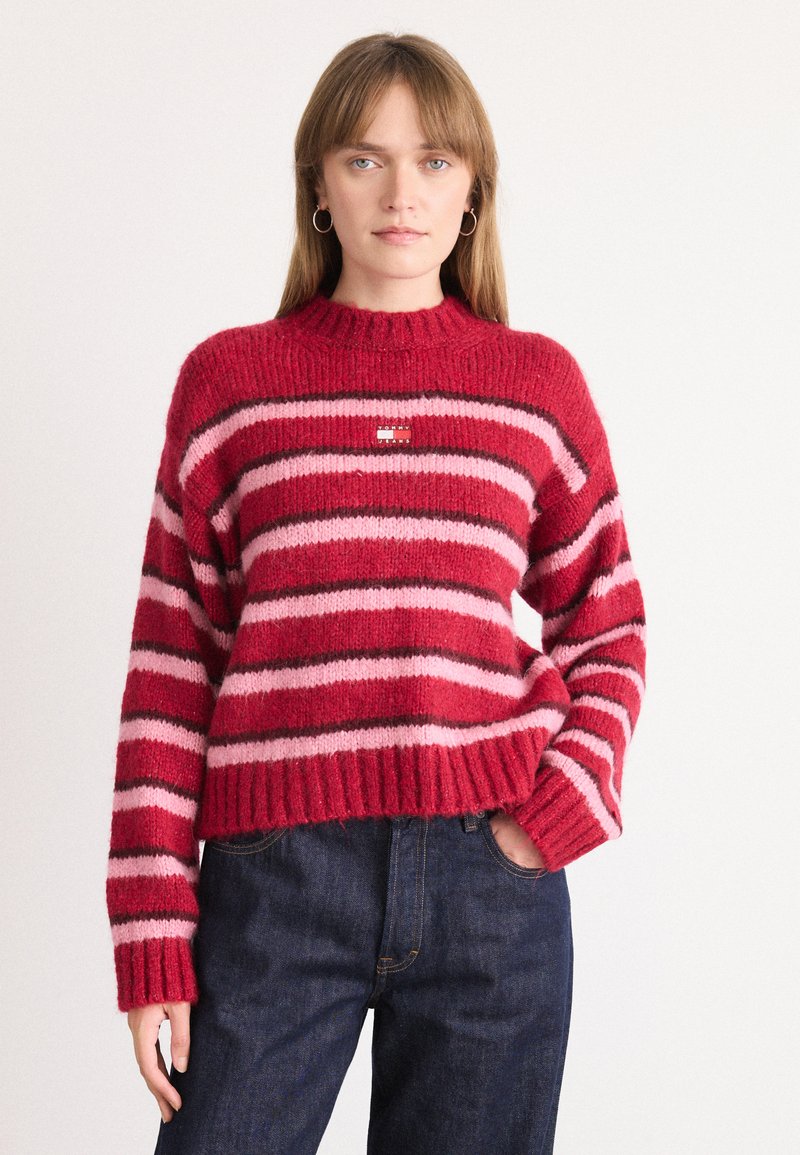 Rood en roze gestreepte trui met een geribde ronde hals en boorden. Zachte textuur, met een klein logo detail aan de voorkant. Gestyled met donkere jeans.