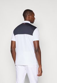 Koszulka polo o białym przodzie i ciemnoniebieskim górnym tyle. Projekt zawiera kołnierzyk oraz czerwone logo na szyi. Krótkie rękawy.