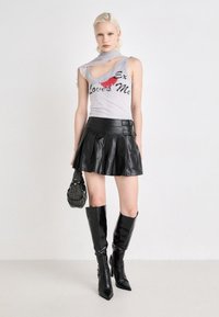 Débardeur gris avec découpe et texte imprimé, associé à une mini-jupe en cuir plissée noire, des bottes noires à talons aiguilles jusqu'aux genoux et un sac en perles.