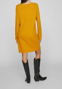 Överdimensionerad senapsgul stickad klänning med långa ärmar, rundad hals, ribbat fåll, i kombination med knähöga svarta läderstövlar.