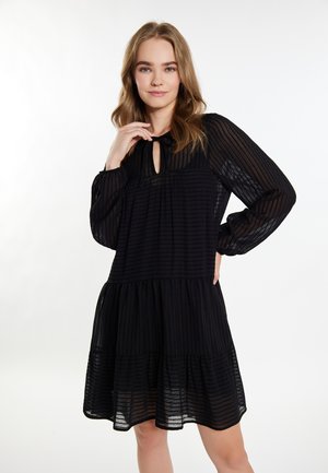 Schwarzes, gestuftes Kleid mit vertikalen Streifen, langen Ärmeln und einem Schlüsselloch-Ausschnitt, aus leichtem Stoff. Hat eine fließende Silhouette.