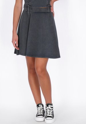 Femeie purtând o fustă cloș de culoare gri închis cu detalii cu șireturi, asortată cu adidași înalți bicolor alb și negru, stând pe un fundal alb.