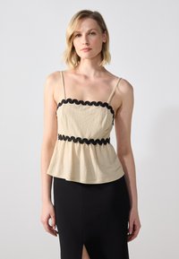 Beige, volangert cami med tynne stropper og svart bølget kant, kombinert med en svart tettsittende skjørt med splitt på siden. Glatt tekstur.