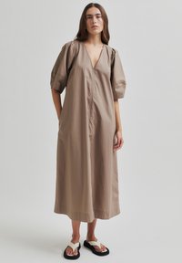 Beige bavlněné šaty s hlubokým výstřihem do V, nabíranými rukávy a rovnou siluetou. Obsahují přední zip a délku do kolen. S kombinací s černými sandály.