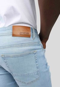 Jeans en denim bleu clair au design ajusté, dotés d'une patch en cuir marron à la taille avec des animaux en relief et de poches arrière standard.