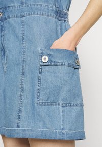 Denim overal v světle modré barvě, který má velkou boční kapsu s detailním knoflíkem a elastický pas pro přiléhavý vzhled.