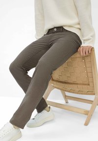 Pantaloni in denim marrone scuro, con texture, a vestibilità slim, dotati di chiusura con bottone frontale e tasche posteriori, abbinati a sneaker bianche.