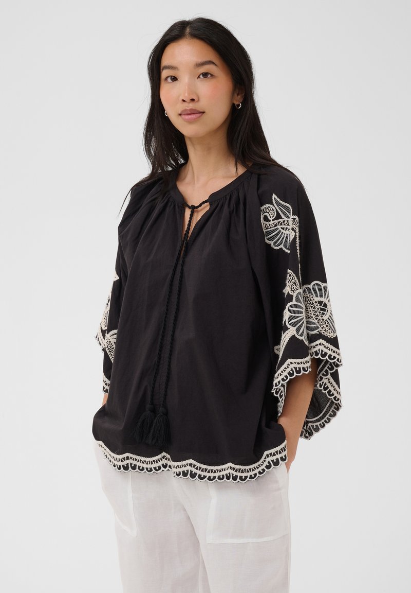 Femme portant un chemisier noir ample avec une broderie florale blanche et des liens à pompons, associée à un pantalon blanc, debout devant un fond uni.