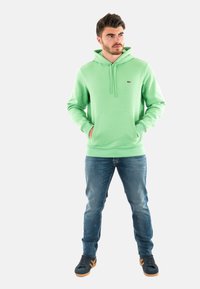 Felpa verde chiaro con tasca frontale e cordini, abbinata a jeans blu e scarpe nere con accenti marroni.
