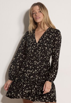 Vrouw met blond haar die een zwarte lange mouwen bloemenjurk draagt met een V-hals en een geplooide taille, staand tegen een lichte achtergrond.