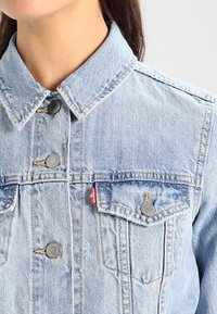 Ljusblå denimjacka med klassisk krage, framknäppning med knappar, två bröstfickor och röd varumärkesetikett.