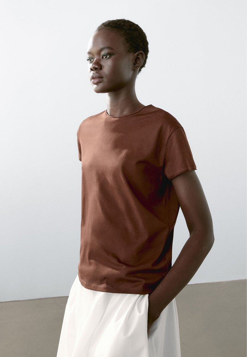 Brun bomulds t-shirt med korte ærmer og rund halsudskæring, kombineret med en hvid stofnederdel. Glat tekstur, afslappet pasform.
