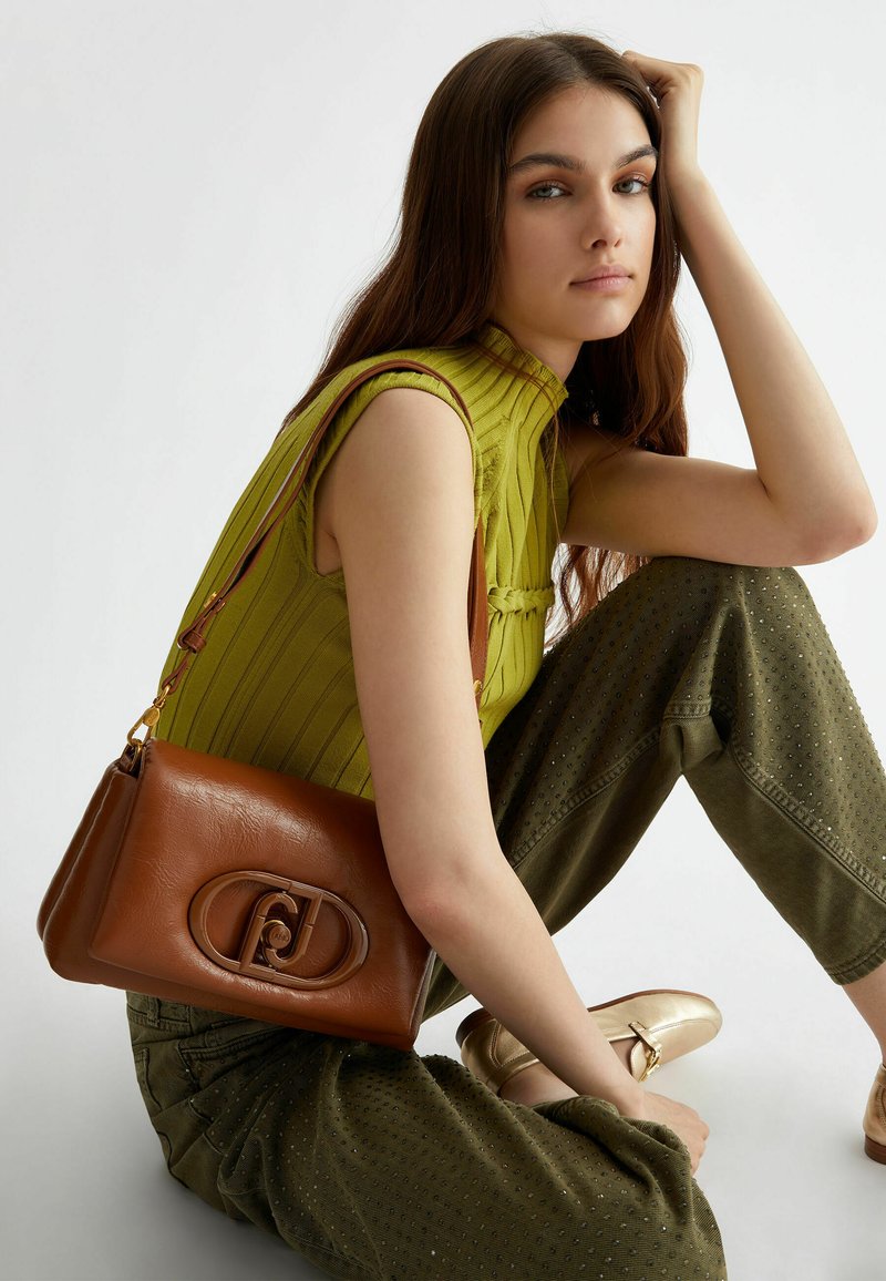 Sac en cuir marron au fini lisse, avec un fermoir décoratif. Porté à l'épaule, associé à un haut vert et à un pantalon olive.