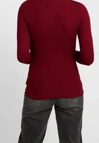 Pull rouge côtelé à manches longues, avec une coupe ajustée et un tissu texturé. Associé à un pantalon ample gris foncé.