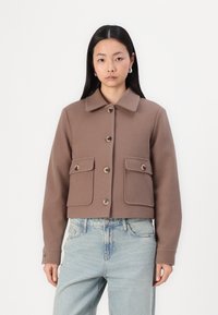 Korte bruine jas van zacht materiaal met een puntige kraag, voorzien van twee voorzakken, knoopsluiting en tonale knopen.