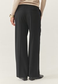 Pantaloni neri a gamba larga con una texture liscia, dotati di due tasche posteriori e una vestibilità su misura. Abbinati a scarpe nere.