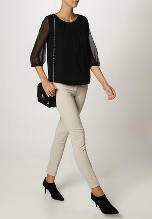 Vrouw die loopt en een zwarte blouse met doorschijnende mouwen draagt, beige smalle broek, zwarte enkellaarsjes met hak, en een kleine zwarte tas met kettingriem draagt.