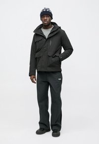 Schwarze Winterjacke mit mehreren Taschen und mehrlagigem Design, kombiniert mit dunkelgrauer Hose und gemustertem Strickmütze.