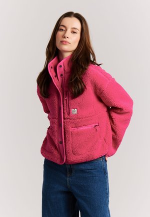 Young woman with long brown hair wearing a bright pink fleece jacket and blue jeans against a plain light background.