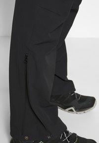 Schwarze Outdoor-Hosen mit Reißverschlüssen an den Säumen, strukturiertem Stoff und geradem Schnitt. Kombiniert mit schwarzen Wanderschuhen mit grauen Akzenten.