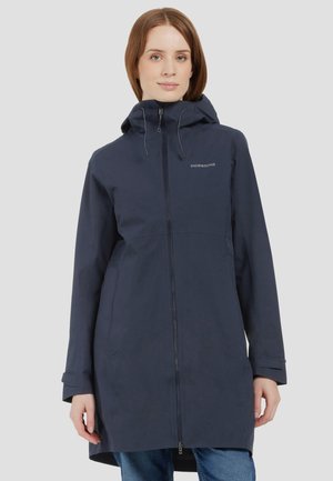 Woman wearing a navy blue waterproof coat with hood and front zipper, standing against a plain light background.