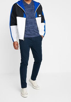 Pánská bunda s barevnými bloky v černé, modré a bílé barvě. Tmavě modré texturované triko s výstřihem do V, tmavě modré kalhoty a bílé tenisky doplňují outfit.