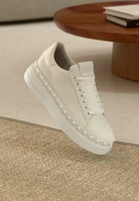 Λευκό δερμάτινο αθλητικό παπούτσι με επίπεδα λευκά κορδόνια, παχιά σόλα και πυραμοειδείς τρουκς κατά μήκος της πλευράς. Λεία υφή, μίνιμαλ σχεδιασμός.