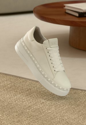 Hvit skinnsneaker med flate hvite lisser, en tykk såle og pyramideformede nagler langs siden. Glatt tekstur, minimalistisk design.