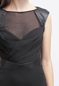 Svart klänning med en genomskinlig mesh-krage, texturerade axelklädslar som liknar läder, och rynkade tygdetaljer över bysten.