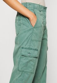 Groene katoenen cargobroek met een losse pasvorm, zijzakken en een knoopaccent. Soepele textuur met zichtbare stikseldetails.
