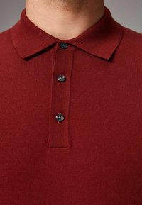 Maroone polo shirt med en tekstureret strik, der har en krave, tre sorte knapper og ribbet detaljering ved stolpen og ærmerne.