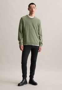 Zielony sweter robiony na drutach z okrągłym dekoltem, noszony na białej bluzce, połączony z czarnymi dżinsami i czarnymi botkami za kostkę. Gładka faktura, luźny krój.