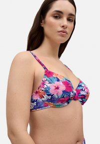 Top de bikini con estampado floral que presenta colores rosa y púrpura, tejido texturizado, tirantes finos y un detalle de nudo en el centro.