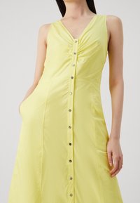Żółta sukienka bez rękawów z dekoltem w kształcie litery V, zgromadzonym biustem i zapięciem na guziki z przodu. Posiada boczne kieszenie i gładką fakturę.