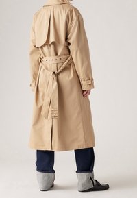 Beżowy płaszcz trench z paskiem w talii, oversize'owym skrzydełkiem na plecach i zapinanymi mankietami. Noszony na ciemnych dżinsach i szarym obuwiu.