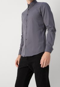 Camisa de manga larga gris con botones y textura suave, que presenta un cuello clásico y un logo en el pecho, combinada con jeans oscuros.