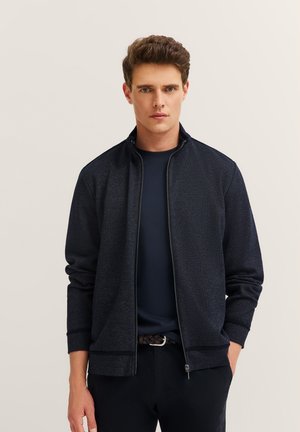 Navy zip-up jacket with a textured fabric, ribbed cuffs, and a relaxed fit, worn over a navy T-shirt, paired with dark trousers.