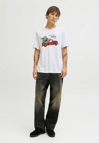 Camiseta de algodón blanca con estampado gráfico de Santa y un personaje verde en un coche rojo. Se lleva con jeans negros deslavados y zapatos negros.