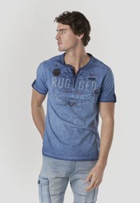 Blaues T-Shirt mit kurzem Arm und Farbverlauf, mit einem knöpfbaren Kragen und einem auffälligen "RUGGED"-Aufdruck sowie verschiedenen Grafiken. Die Textur wirkt weich, legerer Schnitt.