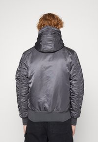 Gray nylon jacket with a hood, featuring a textured finish, elastic cuffs, and ribbed hem. The design includes gathered detailing along the shoulders.