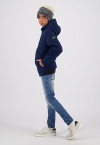 Felpa navy con dettagli verdi, jeans in denim scuro e sneakers bianche. Cappello di maglia con strisce grigie e crema e un logo.