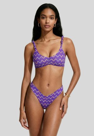 Giovane donna che indossa un bikini viola con motivo a zigzag e spalline arricciate, in piedi su sfondo grigio uniforme, con orecchini a cerchio dorati e bracciale.